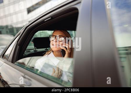 Geschäftsfrau mit Brille, die auf dem Rücksitz des Autos reist und telefoniert, während sie auf das Fenster schaut Stockfoto