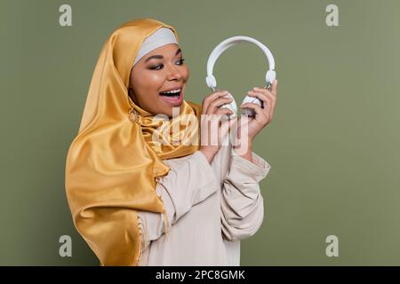 Fröhliche, multirassische Frau in Hijab, die Kopfhörer isoliert auf grünem Stockbild hält Stockfoto