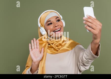 Fröhliche, multiethnische Frau in Hijab und Kopfhörern, die Videoanrufe auf dem Smartphone führt, isoliert auf grünem Stockbild Stockfoto