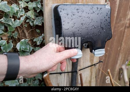 Der Hausbesitzer ist dabei, die wetterfeste Abdeckung einer externen Doppelsteckdose zu öffnen. In einem Garten nach heftigem Regen gesehen Stockfoto