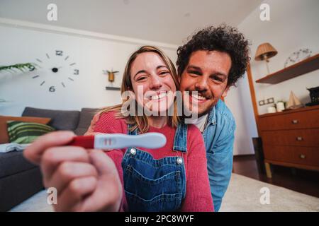 Fröhliches weißes junges Paar, das begeistert lächelt, vor der Kamera ihren positiven Schwangerschaftstest zu Hause zu zeigen. Ehemann und Ehefrau, die feiern, dass sie ein Baby erwarten, das einen Prädiktor in der Hand hält. Hochwertiges Foto Stockfoto