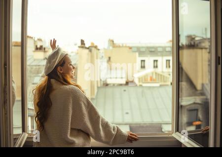 Vor dem Hintergrund der Dächer von Paris steht eine Frau am Fenster. Ein Mädchen mit langen Haaren in einem beigen Anzug und einer Baskenmütze genießt die Stadt und die t Stockfoto