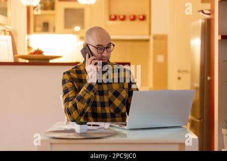 Freiberufliche und Online-Kommunikation. Ein kaukasischer junger Mann mit kahlköpfigem Bart spricht mit dem Handy und arbeitet mit dem Laptop. Konzept von Fernarbeit und Freiberufler. Stockfoto