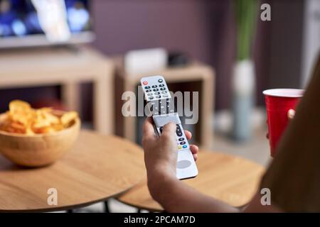 Nahaufnahme eines Mannes, der die Fernbedienung hält, während er zu Hause fernsieht und Snacks genießt, und Platz zum Kopieren Stockfoto