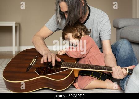 Ein kleines Mädchen sieht sich die Gitarre ihres Großvaters mit Zinsen an, sitzt auf dem Boden. Stockfoto