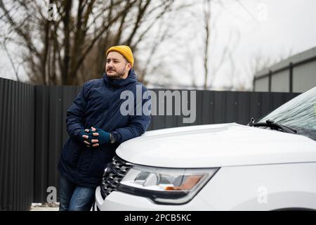 Man-Fahrerjacke und gelber Hut gegen die Motorhaube eines amerikanischen SUV-Wagens bei kaltem Wetter. Stockfoto