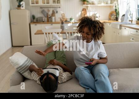 Ein afroamerikanisches Kind spielt zu viele VR-Spiele, die Mutter bittet den Sohn, eine Pause vom 3D-Gaming einzulegen Stockfoto