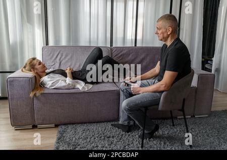 Ein männlicher Psychologe sitzt auf einem Stuhl und spricht mit einem Patienten in einer Psychotherapiesitzung. Positive Erwachsene Frau mit psychischen Problemen liegt auf dem CO Stockfoto