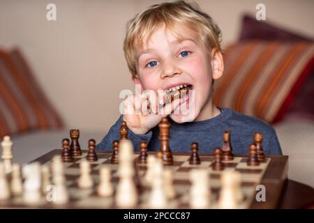 Kleines Kind im Alter von 5 Jahren, das auf einem großen Schachbrett Schach spielt. Schachbrett auf dem Tisch vor dem Jungen, freudig das verlorene Stück essen Stockfoto