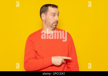 bärtiger hispanischer Mann, der einen roten Pullover trägt, der zur Seite zeigt, wobei der Zeigefinger auf gelbem Studiohintergrund isoliert ist Stockfoto