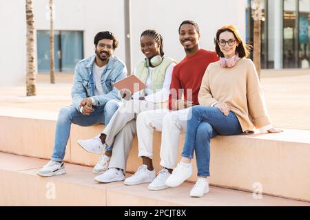 Glückliche junge, multiethnische Schüler, die gelegentlich mit einem Tablet studieren, sich gemeinsam im Freien ausruhen, freien Platz haben Stockfoto