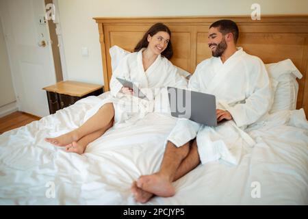 Fröhliches Paar, Das Tablet Und Laptop Benutzt Und Drinnen Auf Dem Bett Sitzt Stockfoto