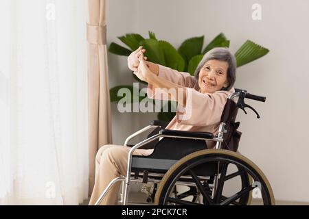 Ältere asiatische Frau, die im Wohnraum zu Hause Stretching-Übungen im Rollstuhl macht Stockfoto