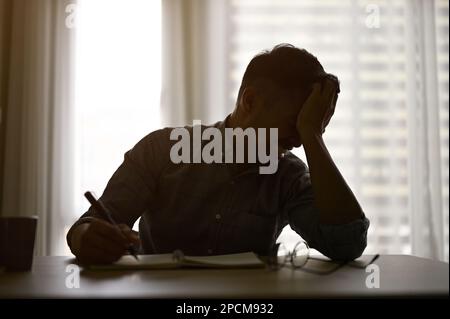 Ein müder und gestresster asiatischer Mann oder Geschäftsmann sitzt an seinem Schreibtisch in einem dunklen Bürozimmer und leidet unter Arbeit, überarbeitet. Silhouettenbild Stockfoto