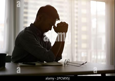 Gestresster und depressiver asiatischer Mann oder Geschäftsmann sitzt am Tisch in einem dunklen Raum. Silhouettenbild Stockfoto