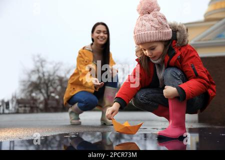 Das kleine Mädchen und ihre Mutter spielen mit einem Papierboot in der Nähe der Pfütze im Freien Stockfoto