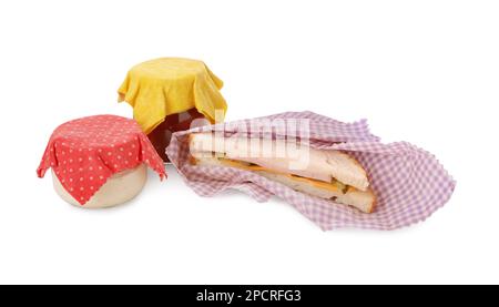 Leckeres Sandwich und Gläser mit Soßen in Bienenwachs-Lebensmittelverpackungen auf weißem Hintergrund Stockfoto
