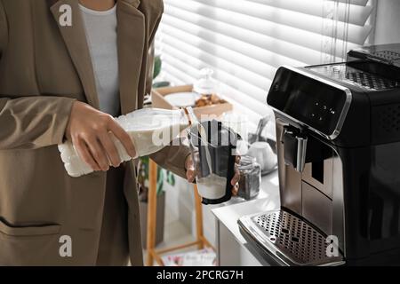 Junge Frau, die Milch in den Behälter neben der modernen Kaffeemaschine im Büro, Nahaufnahme Stockfoto