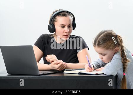 Eine junge Frau mit Kopfhörern arbeitet an einem Laptop und trainiert gleichzeitig mit ihrer Tochter. Stockfoto