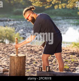 Ein eleganter, brutaler Mann mit Bart und schwarzer Brille hackt Holz auf einem Campingplatz, um ein Feuer anzünden. Outdoor-Aktivitäten im Sommer. Stockfoto