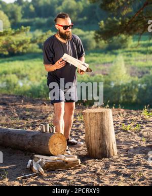 Ein eleganter, brutaler Mann mit Bart und schwarzer Brille hackt Holz auf einem Campingplatz, um ein Feuer anzünden. Outdoor-Aktivitäten im Sommer. Stockfoto