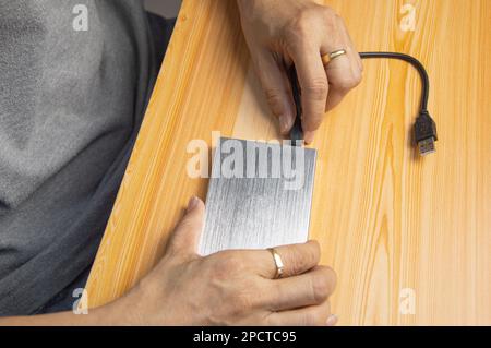 Draufsicht. Mann, der das USB-Kabel einer silbernen externen Festplatte auf dem Holzboden anschließt. Stockfoto