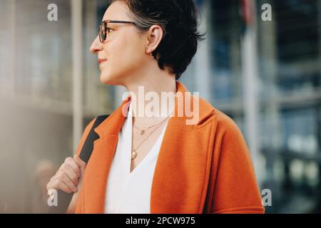 Aufmerksame Geschäftsfrau, die wegblickt, während sie zur Arbeit in der Stadt geht. Reife Geschäftsfrau, die morgens zu Fuß in ihr Büro pendelt. Stockfoto