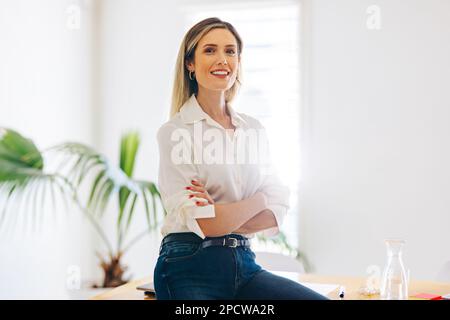 Fröhliche Managerin lächelte in die Kamera, während sie auf einem Konferenztisch saß und die Arme kreuzte. Erfolgreiche Geschäftsfrau, die in einer kreativen Arbeit arbeitet Stockfoto