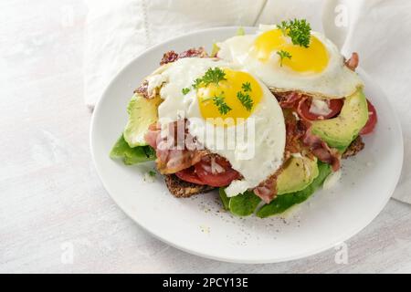 Spiegelei, Speck und Avocado auf dunklem Brot mit Salat und Tomaten, leckeres Frühstückssandwich, heller Hintergrund, Kopierbereich, ausgewählt Stockfoto