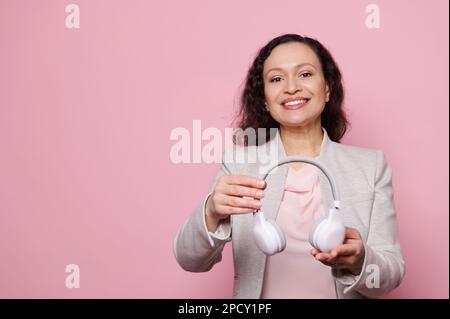 Lächelnde, multiethnische, reife Frau im hellgrauen Anzug, die kabellose Kopfhörer auf einem isolierten pinkfarbenen Hintergrund hält. Speicherplatz kopieren Stockfoto