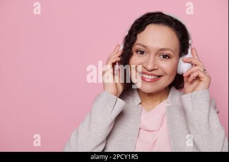 Porträtfoto einer lächelnden hübschen Frau mittleren Alters in kabellosen Kopfhörern, die auf einen isolierten pinken Hintergrund in die Kamera schaut Stockfoto