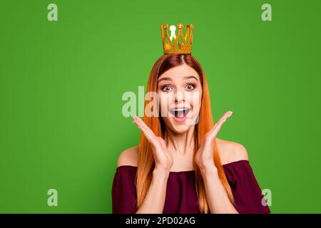 Eine Frau mit dem überraschten Gesicht und der Krone auf dem Kopf öffnete den Mund und die Augen weit und hielt die Hände in der Nähe ihrer Wangen, isoliert auf gelbem Hintergrund Stockfoto