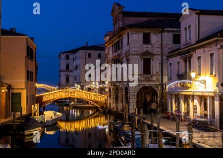 Chioggia Stadtbild in der Lagune von Venedig mit schmalem Vena Wasserkanal mit bunten Booten zwischen alten Gebäuden und beleuchteter Backsteinbrücke am Abend Stockfoto