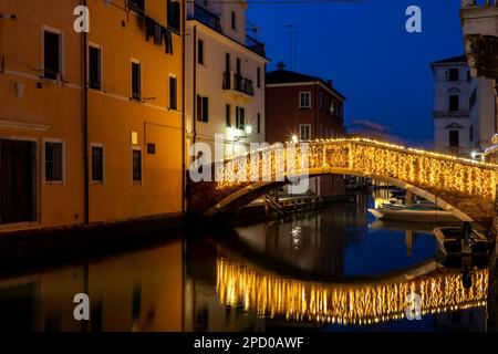 Chioggia Stadtbild in der Lagune von Venedig mit schmalem Vena Wasserkanal mit bunten Booten zwischen alten Gebäuden und beleuchteter Backsteinbrücke am Abend Stockfoto