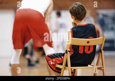 Junger Basketballspieler, Der Basketball-Übungsübungen Anschaut. Ein Junge sitzt auf einem Holzstuhl und ruht sich während des Basketballtrainings aus. Basketball-Camp Stockfoto