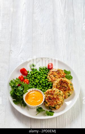 Krabbenkuchen mit Remouladesauce, grünen Erbsen, Tomaten, frischem Koriander auf weißem Teller auf weißem Holztisch, vertikale Sicht von oben, freier Platz Stockfoto