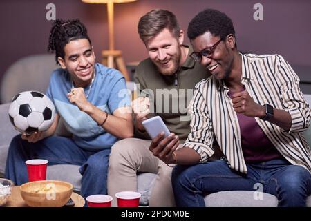 Eine vielfältige Gruppe von Freunden, die sich ein Sportereignis online über ihr Smartphone ansehen und emotional anfeuern Stockfoto
