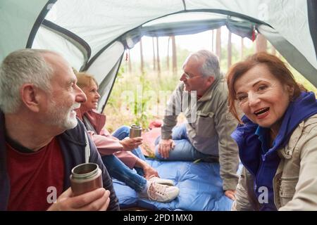 Glückliche Seniorin, die während des Urlaubs bei männlichen und weiblichen Freunden im Zelt sitzt Stockfoto