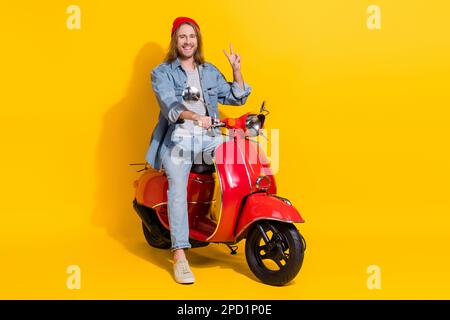 Foto in voller Länge von einem coolen Mann, der eine Jeansjacke auf einem Moped trägt und auf dem ein V-Schild in Gelb angezeigt wird Stockfoto