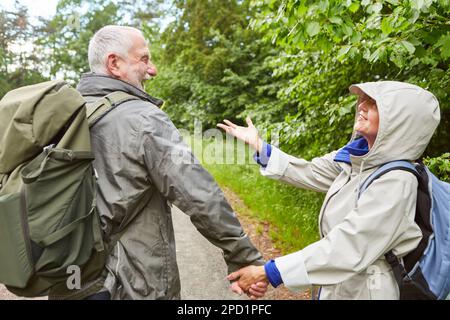 Fröhliches älteres Paar mit Rucksäcken und Jacken, die während des Urlaubs beim Wandern im Wald Händchen halten Stockfoto