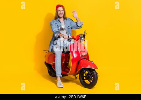 Langes Foto eines coolen, freundlichen Mannes, der eine Jeansjacke trägt, die mit den Armen auf einem isolierten gelben Hintergrund mit Moped herumwedelt Stockfoto