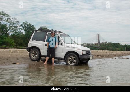 Junger Tourist neben seinem Geländefahrzeug am Rand eines Flusses im Amazonas-Dschungel während eines bewölkten Tages Stockfoto