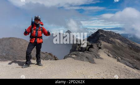 Junger Kletterer auf dem Gipfel des Vulkans Guagua Pichincha, der an einem bewölkten Tag rote Jacke und Helm trägt Stockfoto