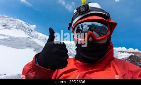 Kletterer mit Helm, Stirnlampe und schwarzer Jacke macht ein Selfie auf einem Gletscher an einem bewölkten Tag, die Landschaft spiegelt sich in rot gerahmten Gläsern wider Stockfoto