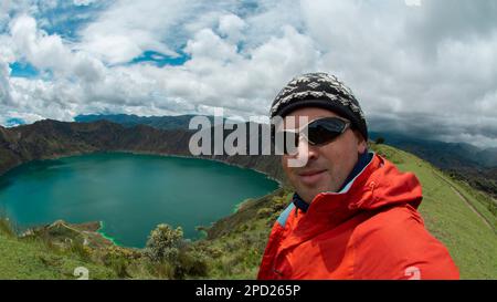 Junger Tourist in rotem Mantel und Gläsern, der ein Selfie in der Nähe des Kraters des Vulkans Quilotoa in Ecuador macht Stockfoto