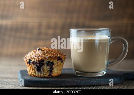 Köstlicher Muffin mit Blaubeeren und Cappuccino-Glasbecher auf einem Holztisch, Nahaufnahme. Frischer Cupcake und Kaffee zum Frühstück, Kopierbereich Stockfoto