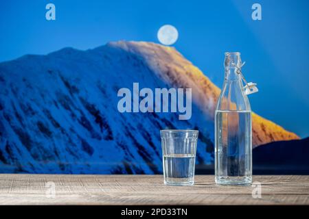 Eine Flasche reines Wasser und ein Glas auf einem Holztisch mit dem schneebedeckten Hintergrund der Himalaya Berge und Vollmond, aus nächster Nähe Stockfoto