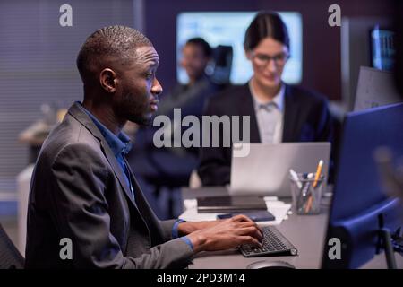 Seitliches Porträt eines seriösen schwarzen Geschäftsmanns, der am Arbeitsplatz in einem High-Tech-IT-Sicherheitsbüro ein Notebook verwendet Stockfoto