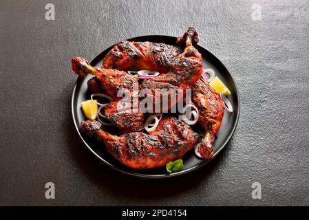 Tandoori-Hühnchen im indischen Stil auf dem Teller mit dunklem Steinhintergrund. Hähnchenschenkel mariniert in Joghurt und Gewürzen. Nahaufnahme Stockfoto