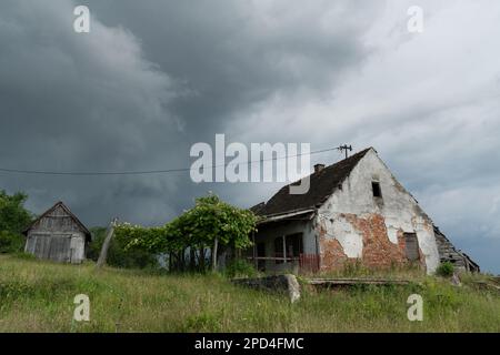 Sturmwolken über einem verlassenen Landhaus, dunkle, dramatische Wolken über leeren, verlassenen Ruinen, die ein Haus mit einem Garten überwuchert im Gras ruinieren Stockfoto